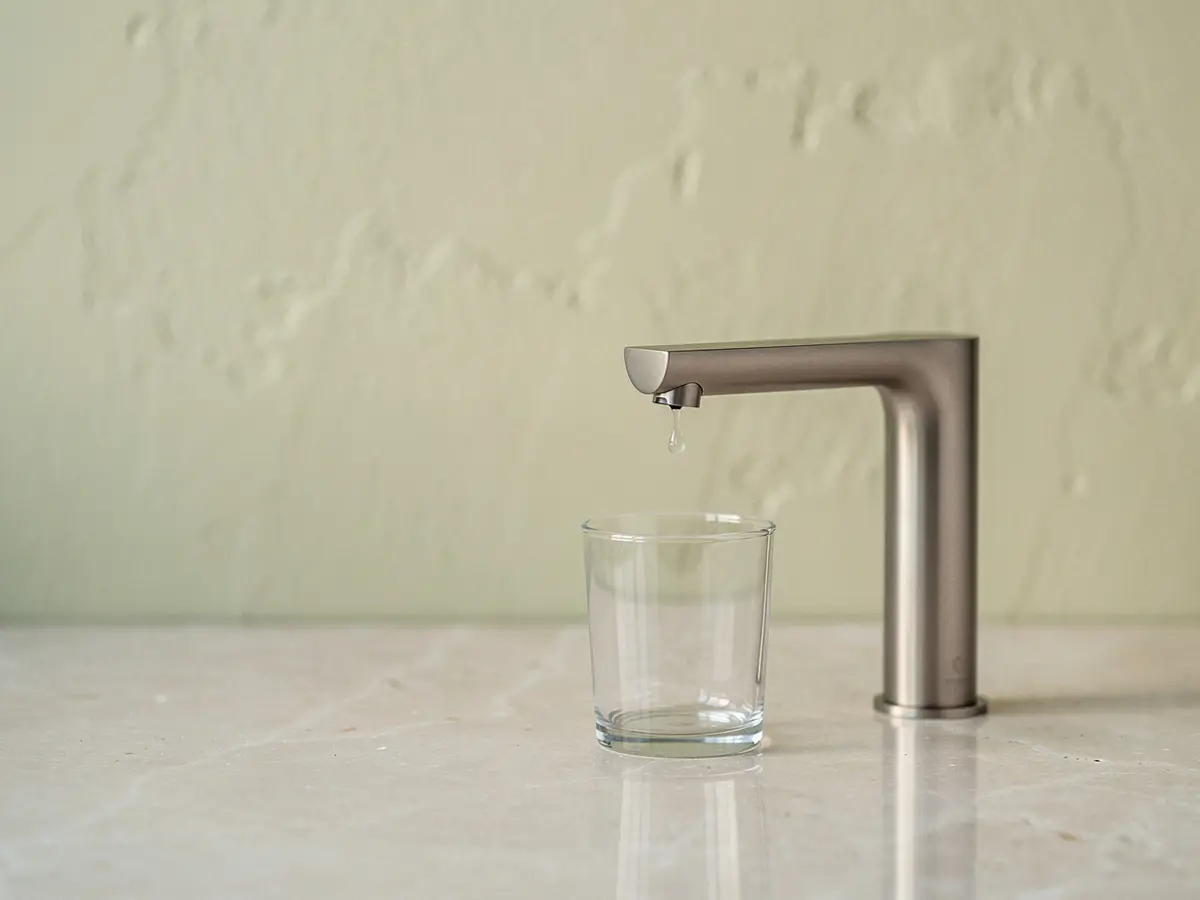 Gota de agua formándose en el caño de un grifo de cocina, con un vaso debajo (imagen generada con IA)